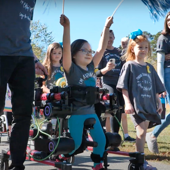 A child using robotic gait trainers to walk alongside other children and parents while holding and raising two pom-poms in her hands.